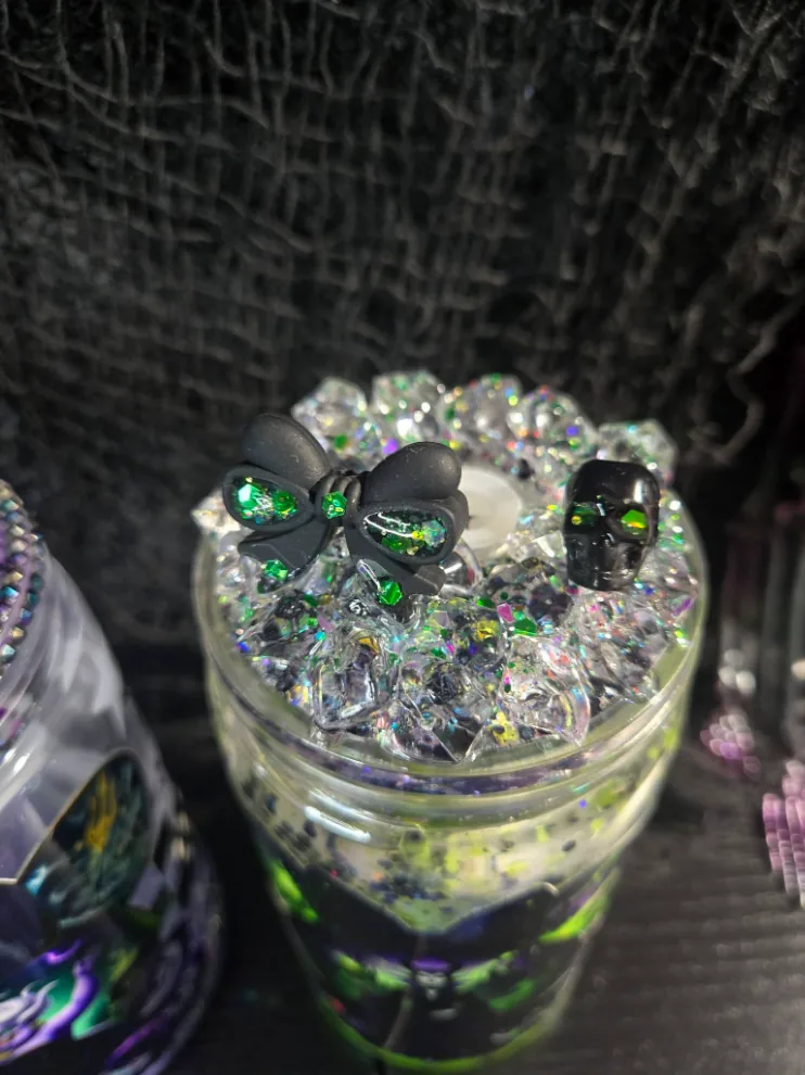 Close-up of a tumbler lid piled with clear crushed crystal and green glitter, topped with a black bow charm and a black skull with green gem eyes.