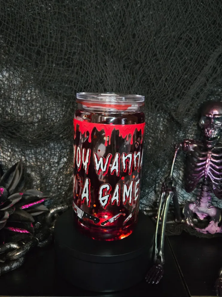 Blood-red glitter tumbler with a dripping red band and white horror lettering reading You wanna play a game, set beside a purple skeleton.