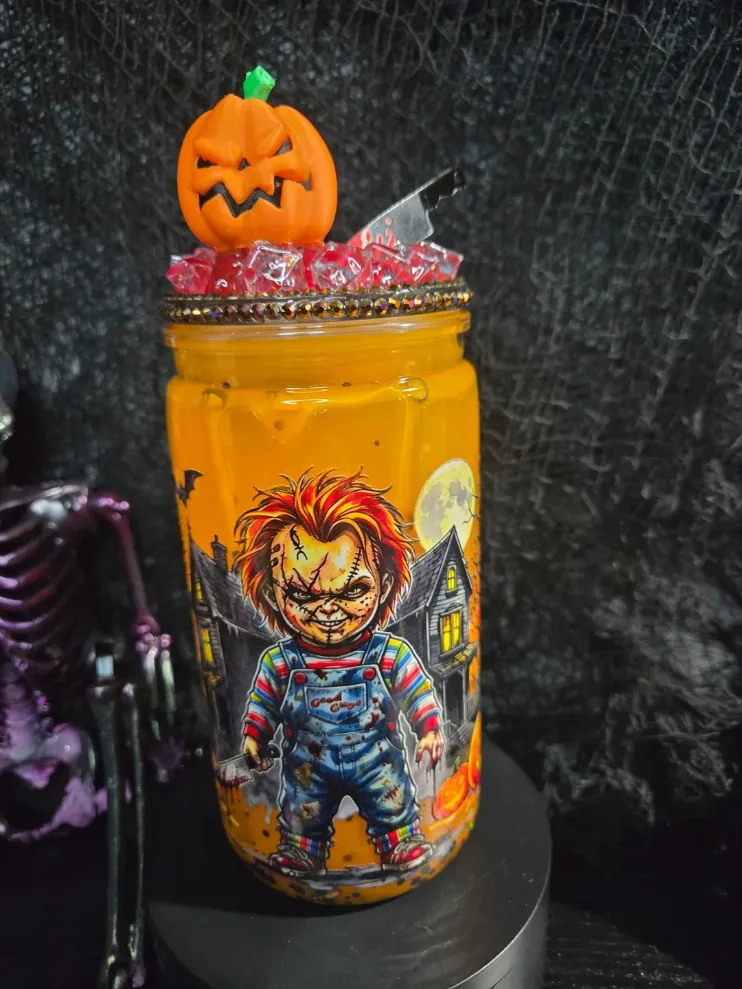 Orange glitter tumbler with a horror doll print in front of a haunted house, finished with a rhinestone rim, red crystal lid, a jack-o’-lantern topper and a tiny cleaver.
